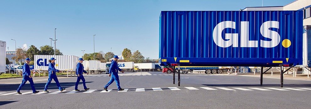 Équipe GLS sur site logistique traversant une zone sécurisée devant une semi-remorque bleue, centre de distribution et transport de colis.