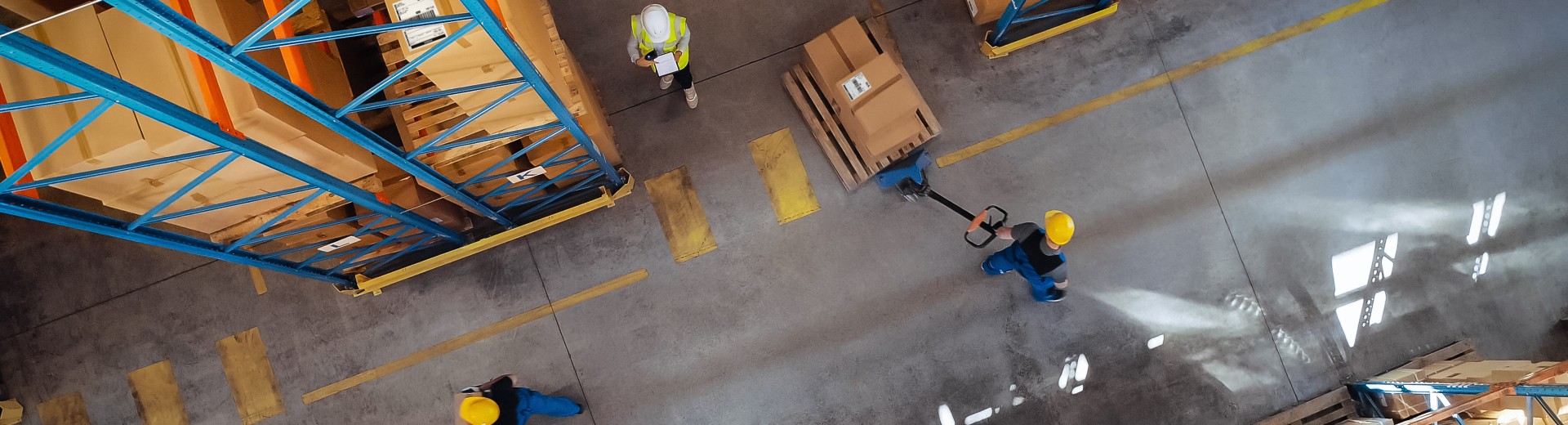 Overhead view of three warehouse workers performing separate tasks among aisles of stacked boxes.